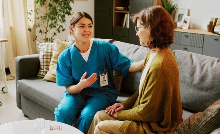 Healthcare worker sitting beside elderly lady in cozy living room providing comfort and care, indoor setting with natural light filtering through windows